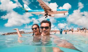 a woman and man sitting in the sea at maho beach with a plane flying over them